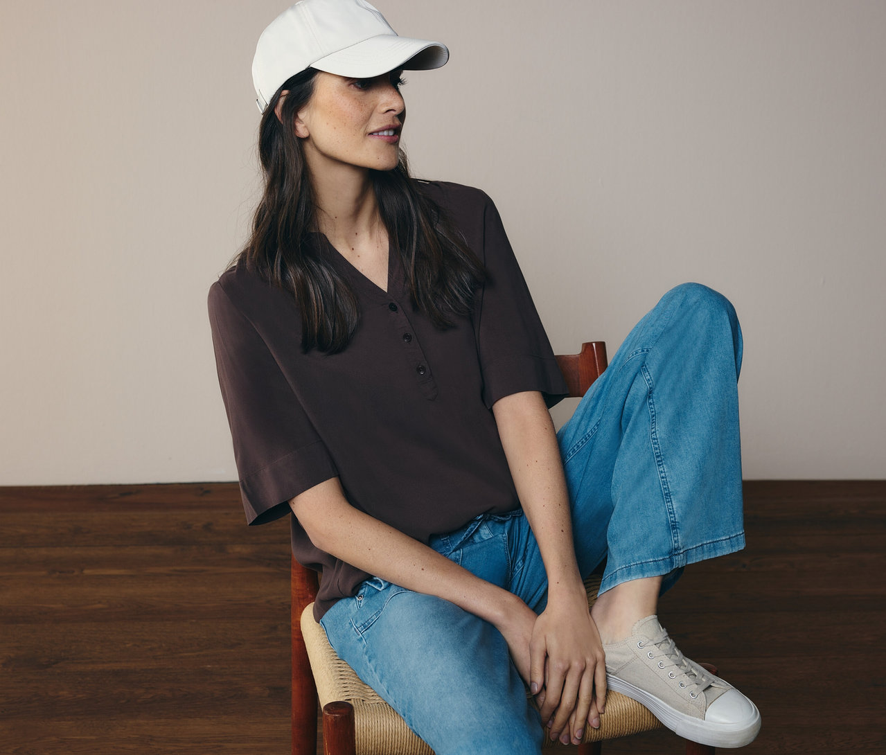 Žena sedí na židli, má džíny, čepici a hnědou tunikovou halenku CECIL.