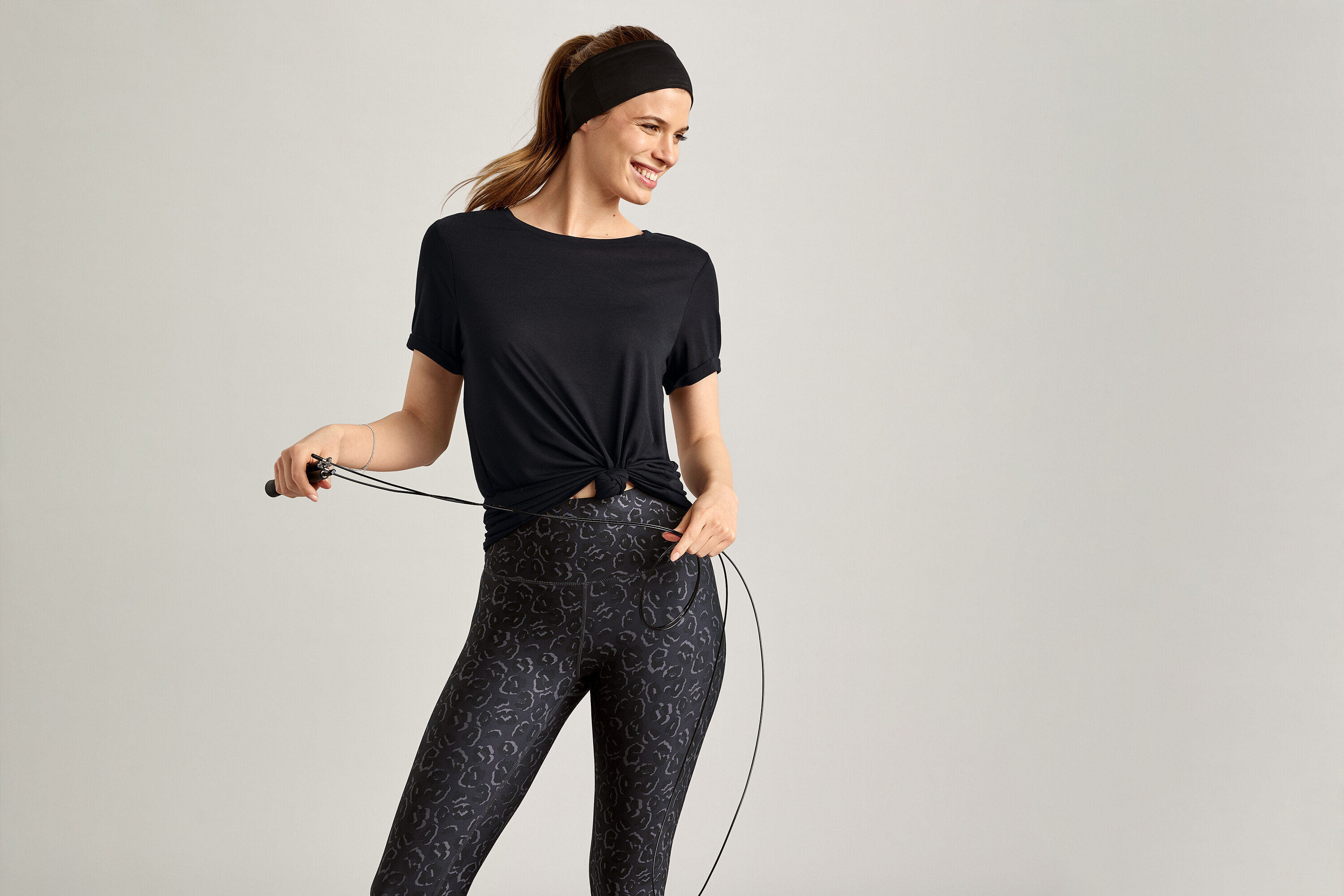 Žena s čelenkou a v černém sportovním tričku drží švihadlo na šedém pozadí.
