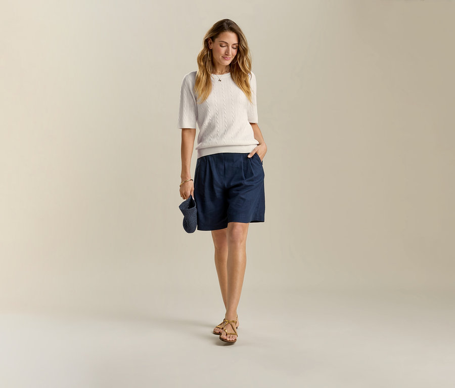 Žena v tmavě modrých lněných bermudách a sandálech. V ruce drží tmavě modrou čepici.