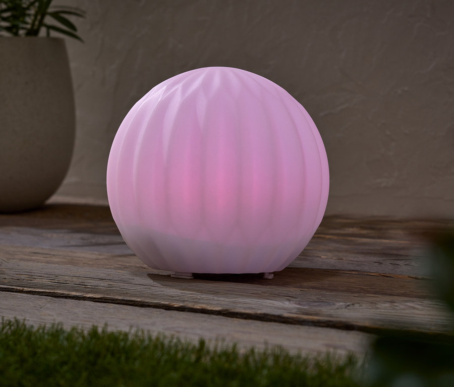 Růžová kulová lampa stojí na dřevěném stole vedle květináče s rostlinou.