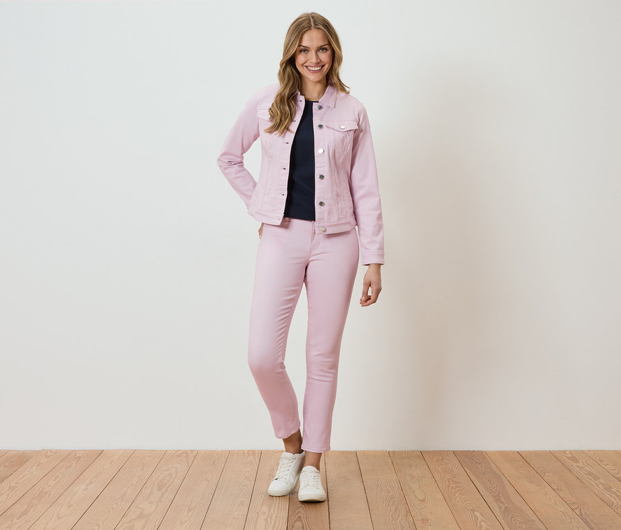 Žena stojí před stěnou a má na sobě růžovou džínovou bundu a kalhoty.