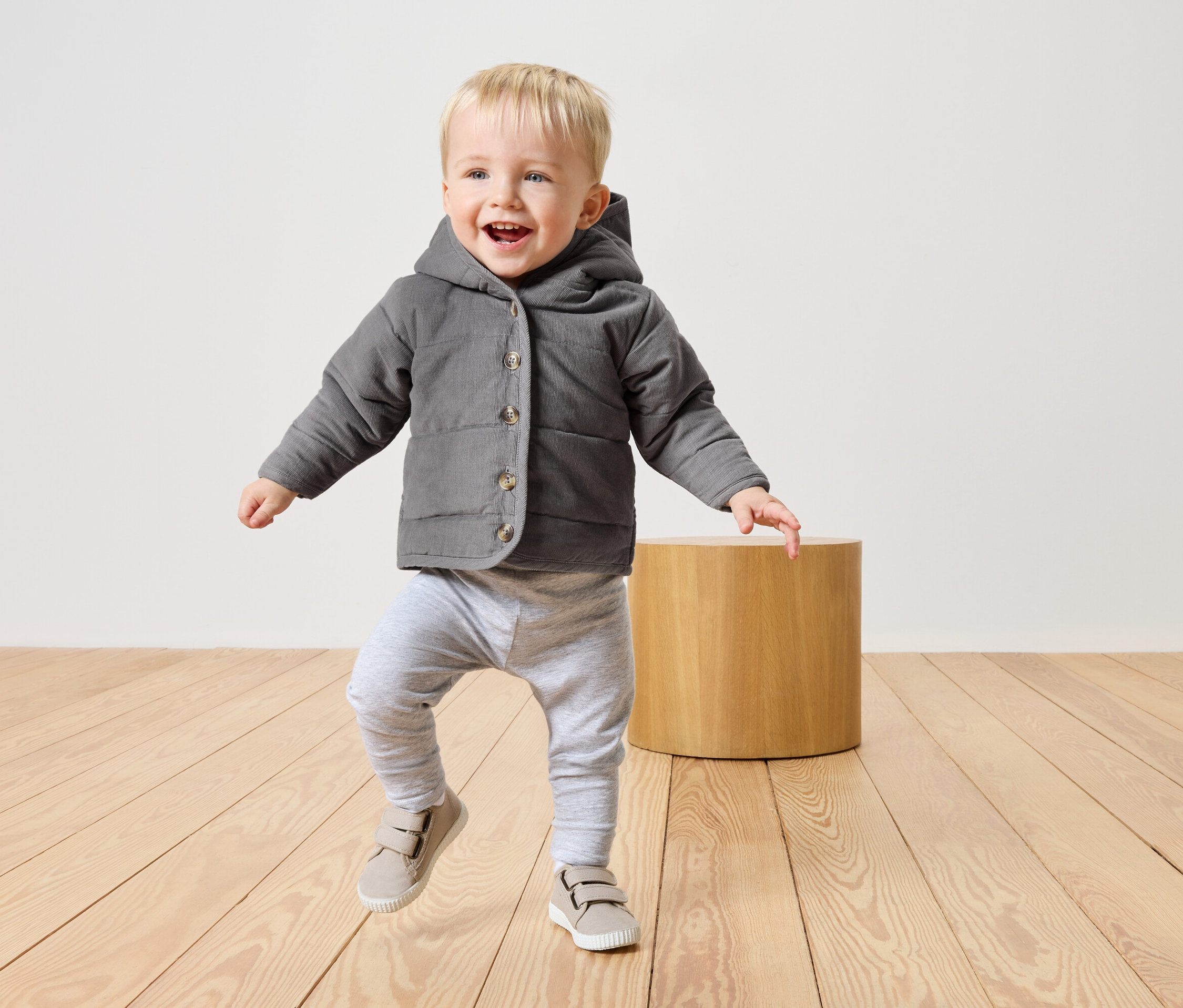 Malý chlapec s blond vlasy v šedé bundě s kapucí, šedých kalhotách a béžových botách běží po světlé dřevěné podlaze.