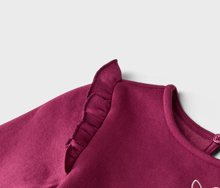 Detail bordové mikiny s volánky na rameni a knoflíkem na krku.