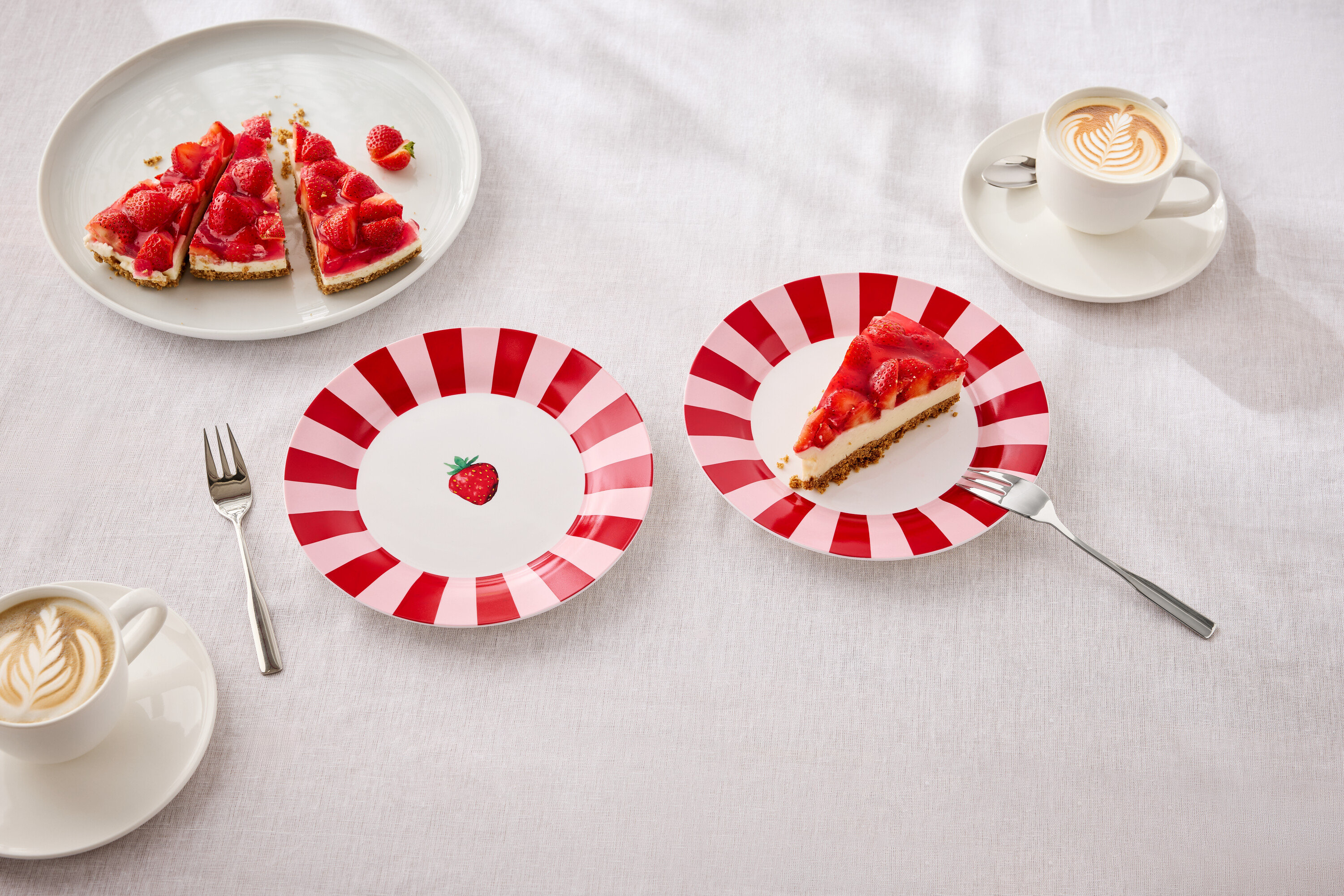 Na stole jsou talíře s jahodovým koláčem, vidličky a šálky s latte art.