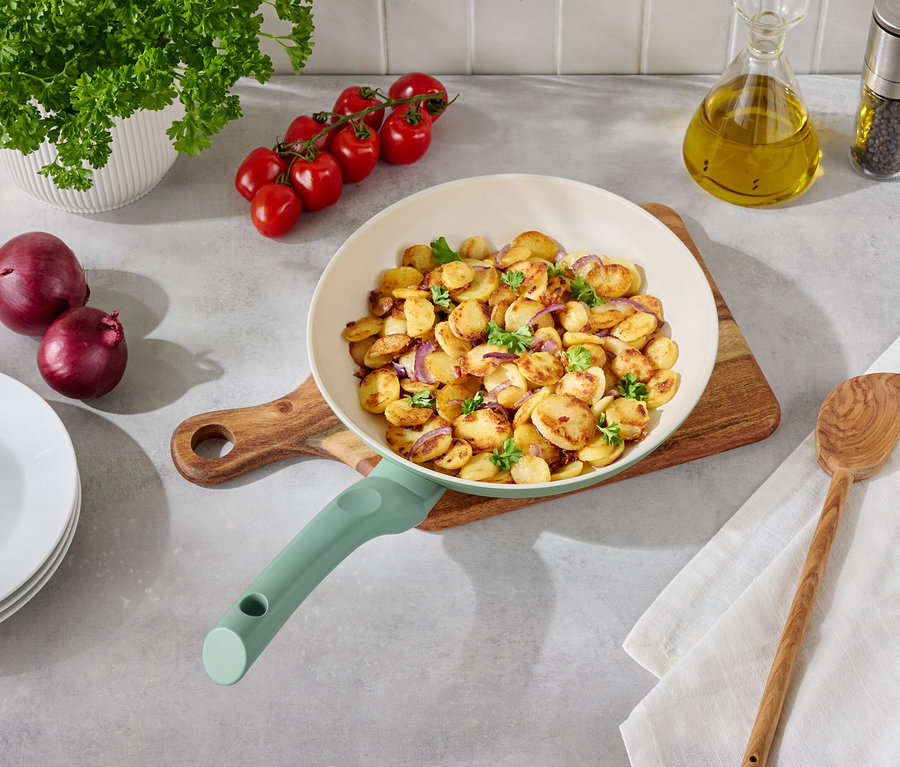 Smažené brambory s cibulí a petrželkou na pánvi na dřevěném stole s přílohami.