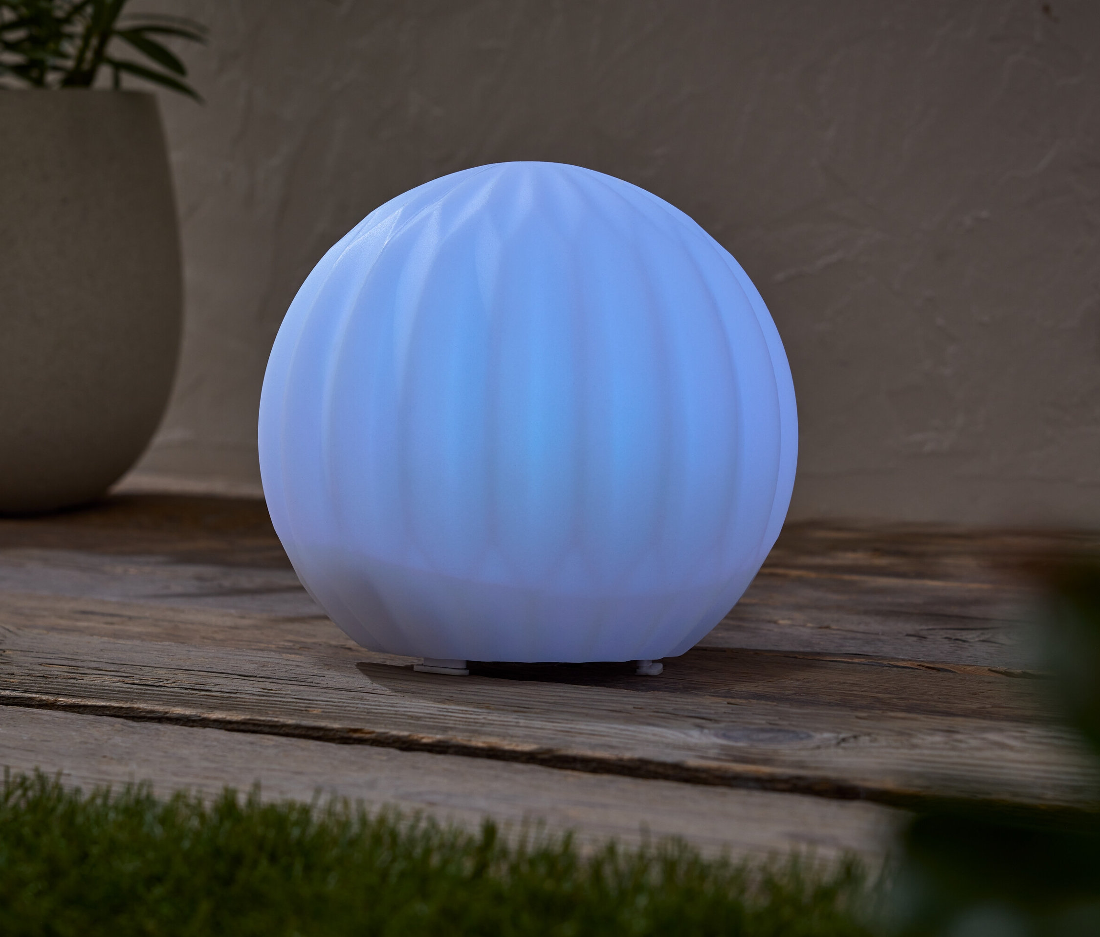 Svítící kulovitá lampa stojí na dřevěném stole vedle květináče s rostlinou.