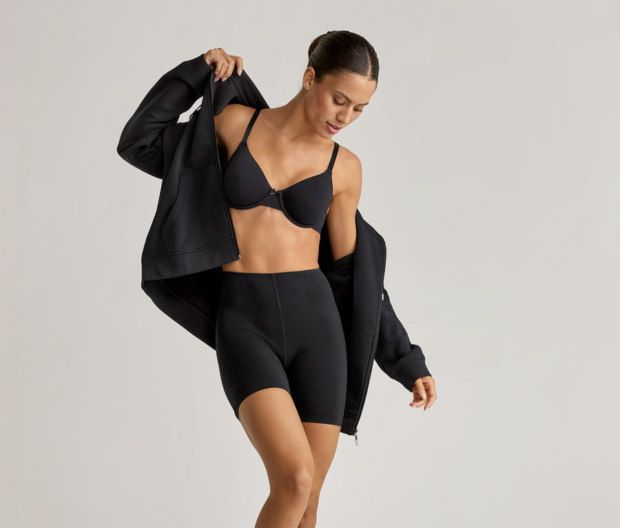 Žena v černé podprsence s kosticemi, cyklistických šortkách (2 ks) a pohodlné mikině, kterou si rozepíná.