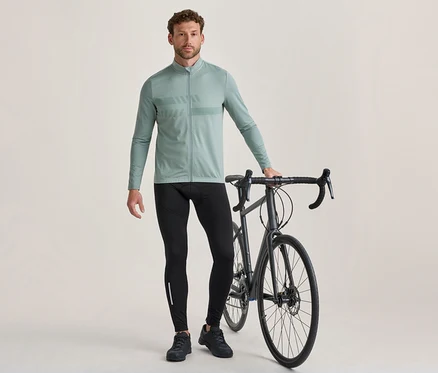 Muž stojí vedle tmavě šedého silničního kola a drží jeho řídítka. Má na sobě světle zelený cyklistický dres s dlouhým rukávem a vzorem na hrudi, černé cyklistické legíny s reflexním pruhem a černé cyklistické boty.