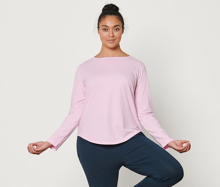 Žena stojí na jógové podložce v pozici stromu. Má na sobě světle růžové sportovní triko s dlouhým rukávem a kalhoty na jógu.