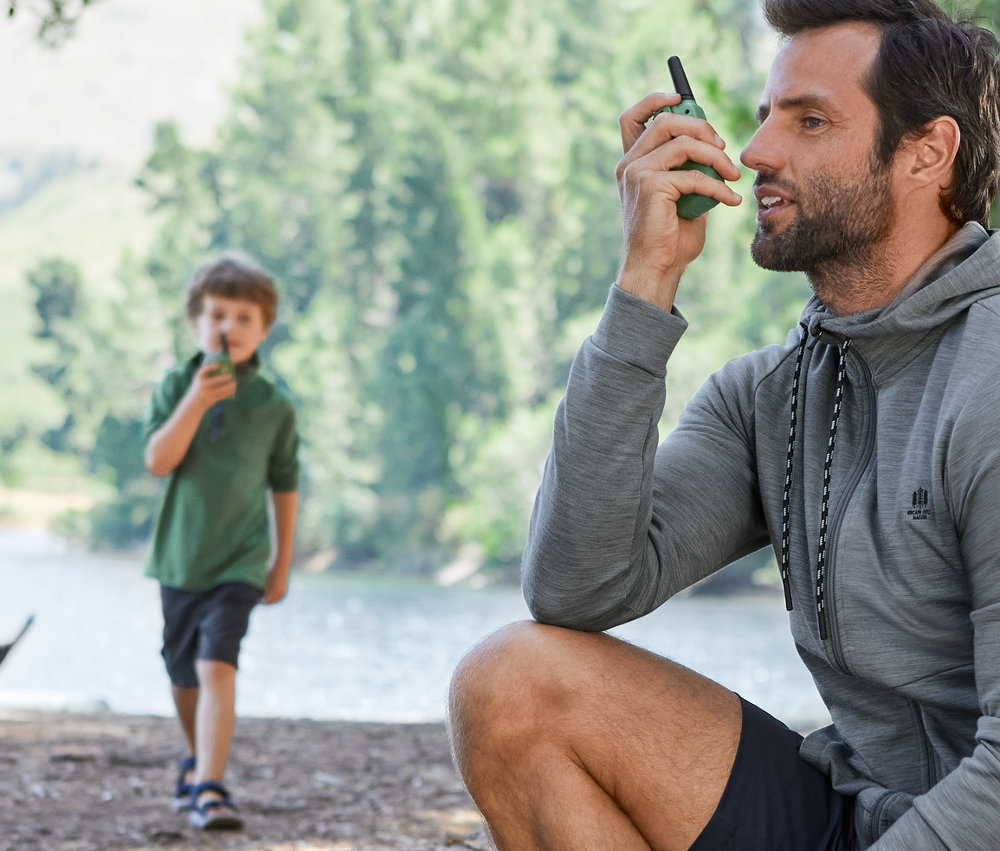 Muž sedí na kládě a používá vysílačku. V pozadí je chlapec s vysílačkou. Oba jsou oblečeni ve funkčním oblečení.
