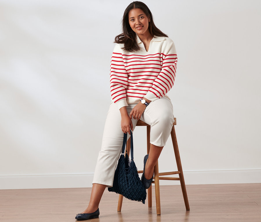 Žena sedí na dřevěné stoličce a má na sobě bílo-červený svetr s límečkem, džíny slim fit, kožené baleríny a náramkové hodinky z ušlechtilé oceli. V ruce drží síťovku.