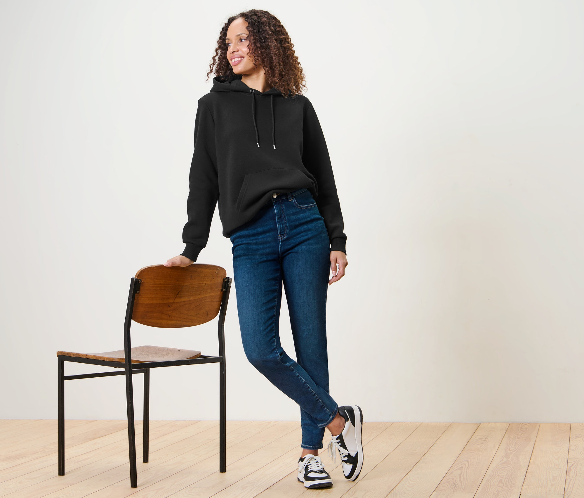 Žena v černé mikině a modrých džínách »Hanna« stojí opřená o židli.