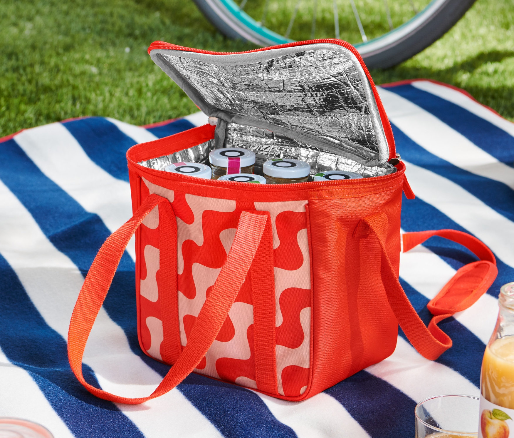 Otevřená malá chladicí taška s červeno-bílým vzorem stojí na dece s modro-bílými pruhy. Uvnitř jsou vidět sklenice. Kolo a helma leží v pozadí.