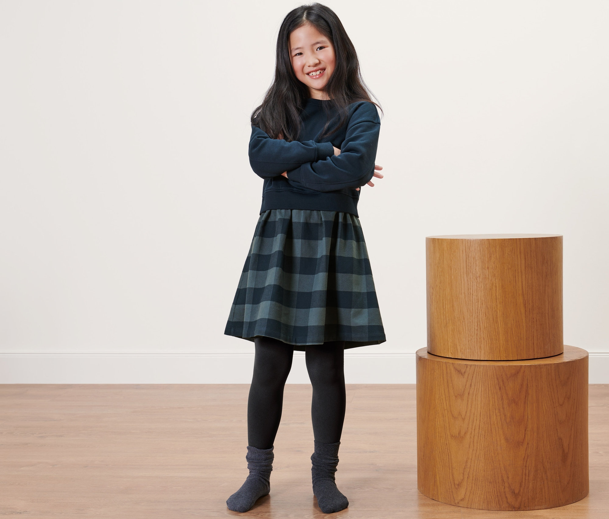 Usmívající se dívka pózuje s rukama zkříženýma na sobě a má na sobě dětské termo legíny, svetřík a kostkovanou sukni.