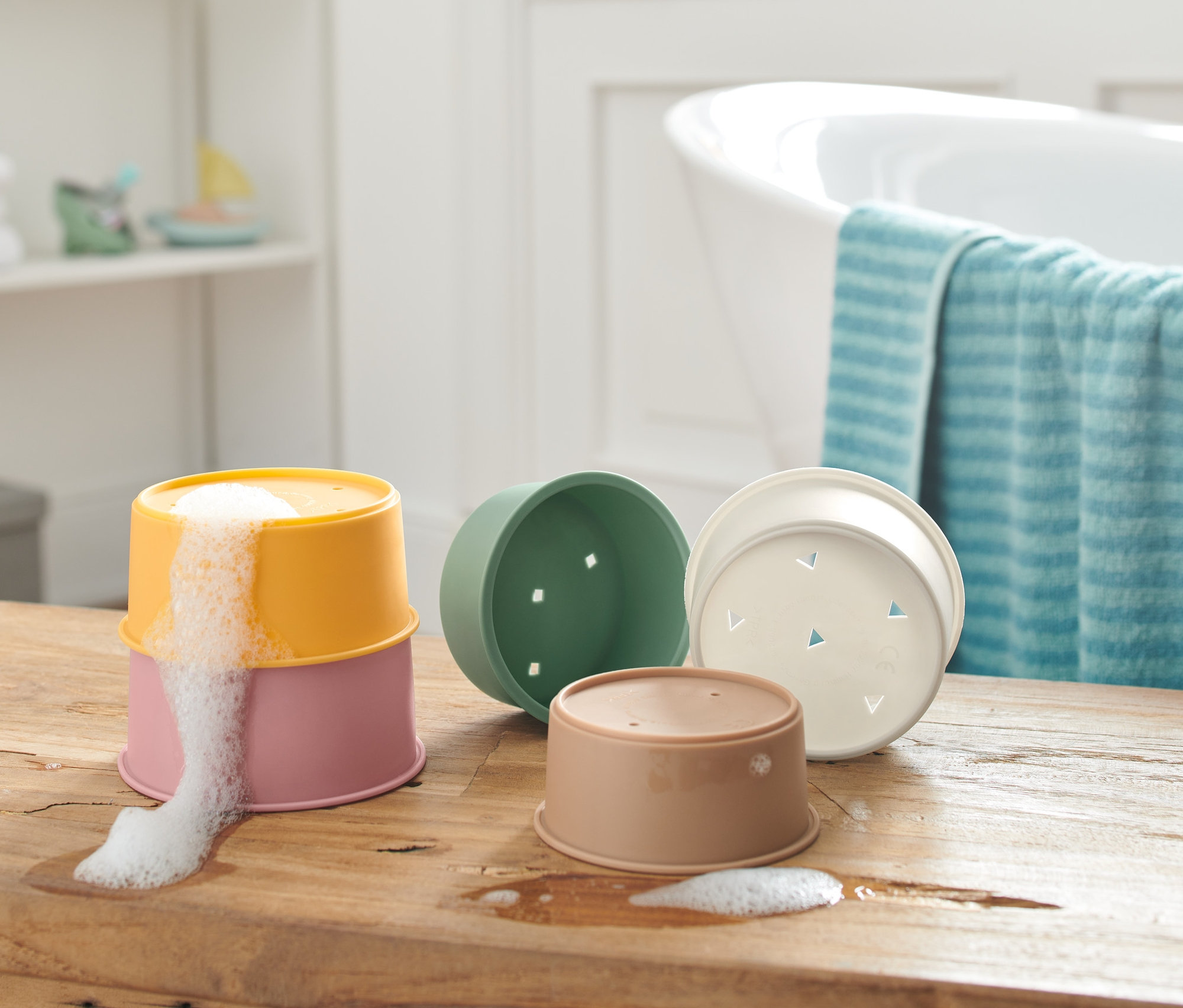 Na dřevěném stole jsou stohovatelné kelímky na hraní do vody, dva na sobě s pěnou vytékající z horního kelímku.