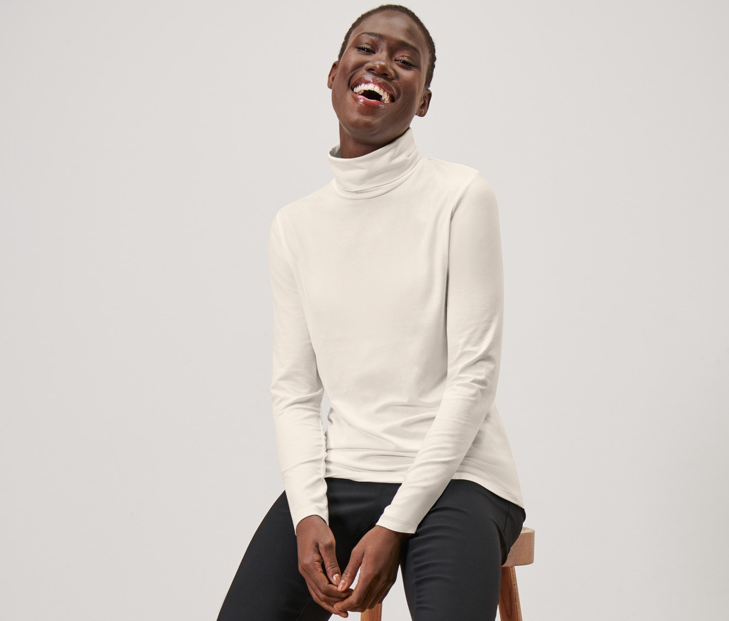 Žena sedí na stoličce a má na sobě krémově bílé triko s rolákem, černé kalhoty a černé mokasíny.