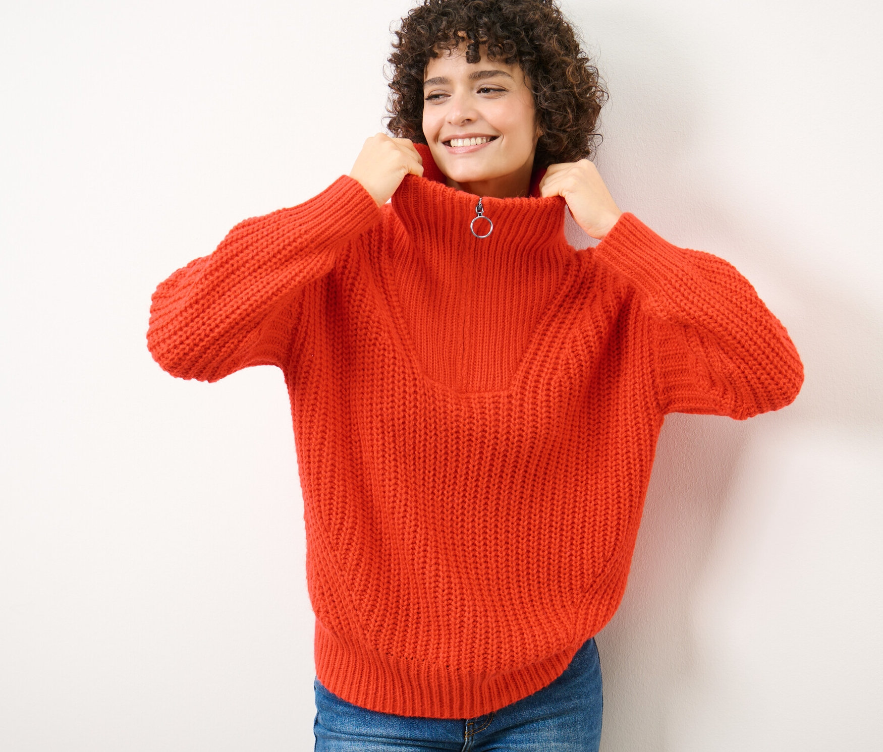 Žena si přitahuje límec oranžového svetru troyer z hrubého úpletu ke krku.