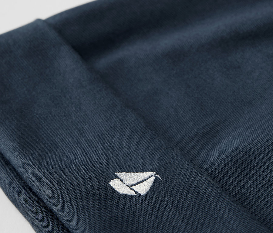 Detail modré látkové čepice ze soupravy pro miminka s vyšitým bílým logem.