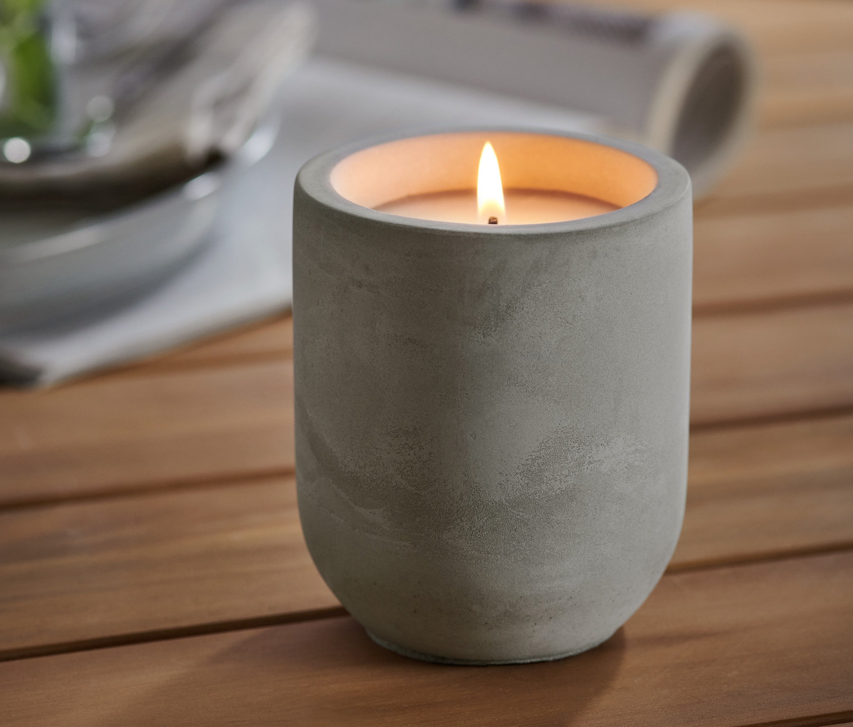 Na dřevěném stole stojí zapálená citronelová svíčka v cementovém obalu. V pozadí je sklenice s rostlinou a talíře.