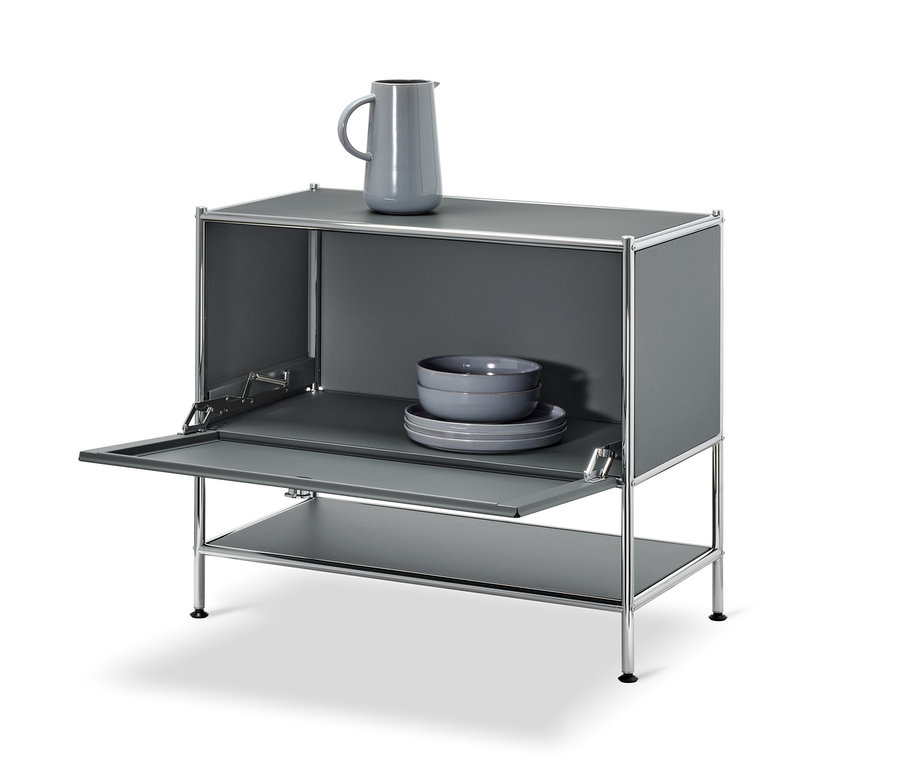 Šedá odkládací skříňka CN3 s otevřenými výklopnými dvířky. Na horní části skříňky je šedá váza, uvnitř jsou šedé misky a talíře.