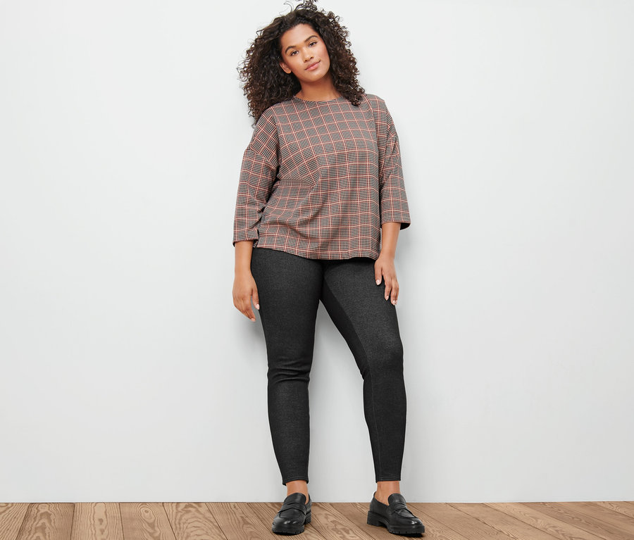 Žena pózuje v mikině s 3/4 rukávem se vzorem kohoutí stopy a v džeginách.