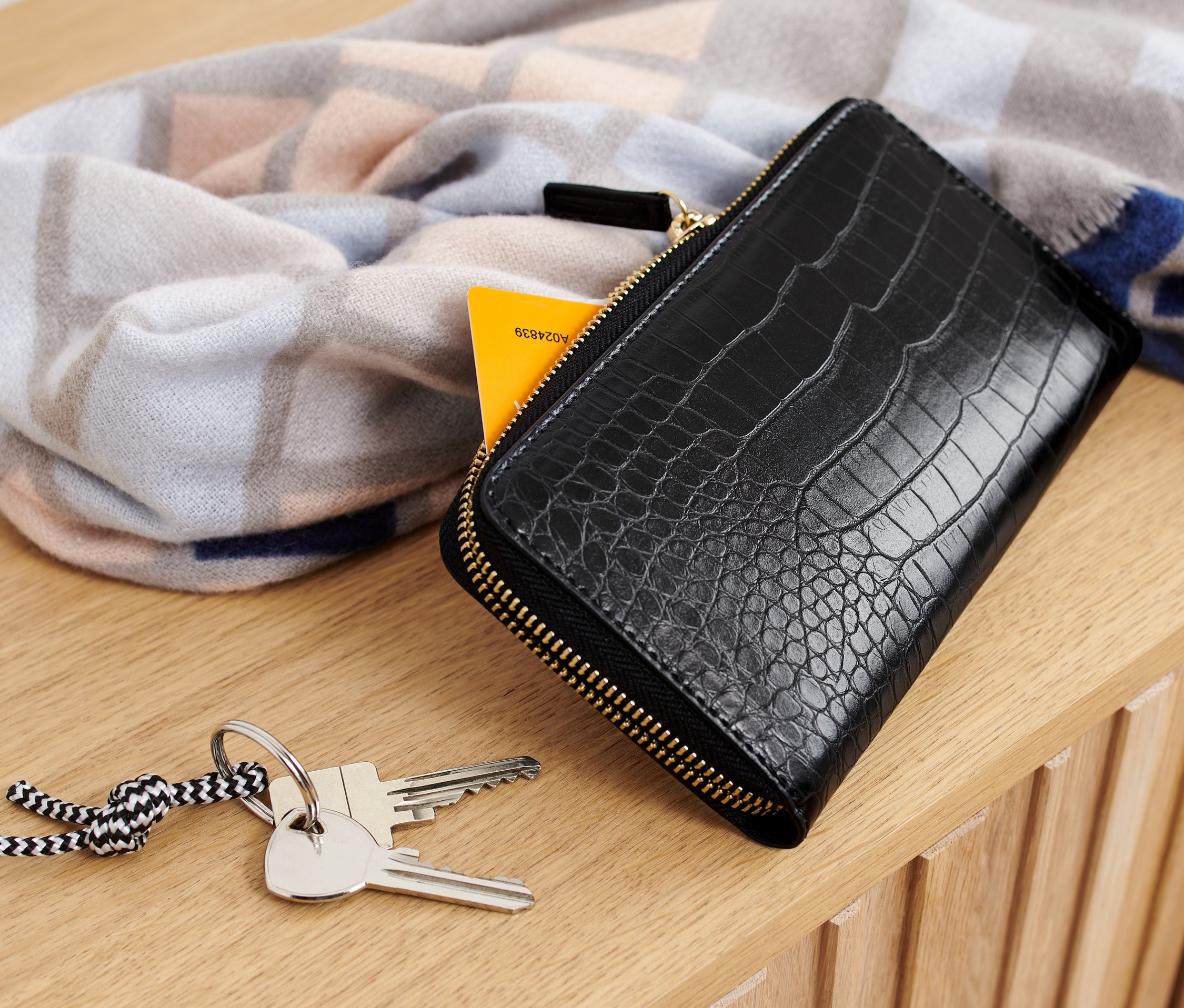 Černá peněženka leží na dřevěném povrchu vedle klíčů a vzorované šály.