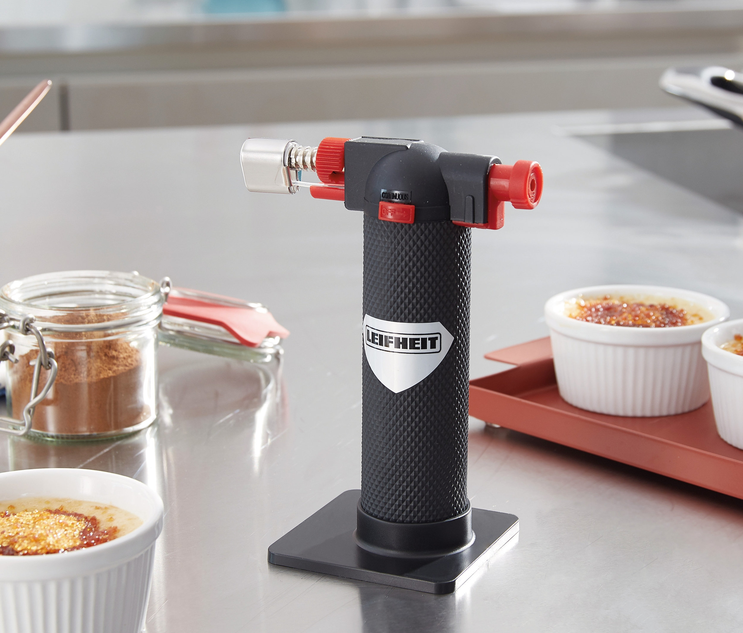 Na stole stojí hořák Gourmet a flambovací pistole vedle misek s crème brûlée.