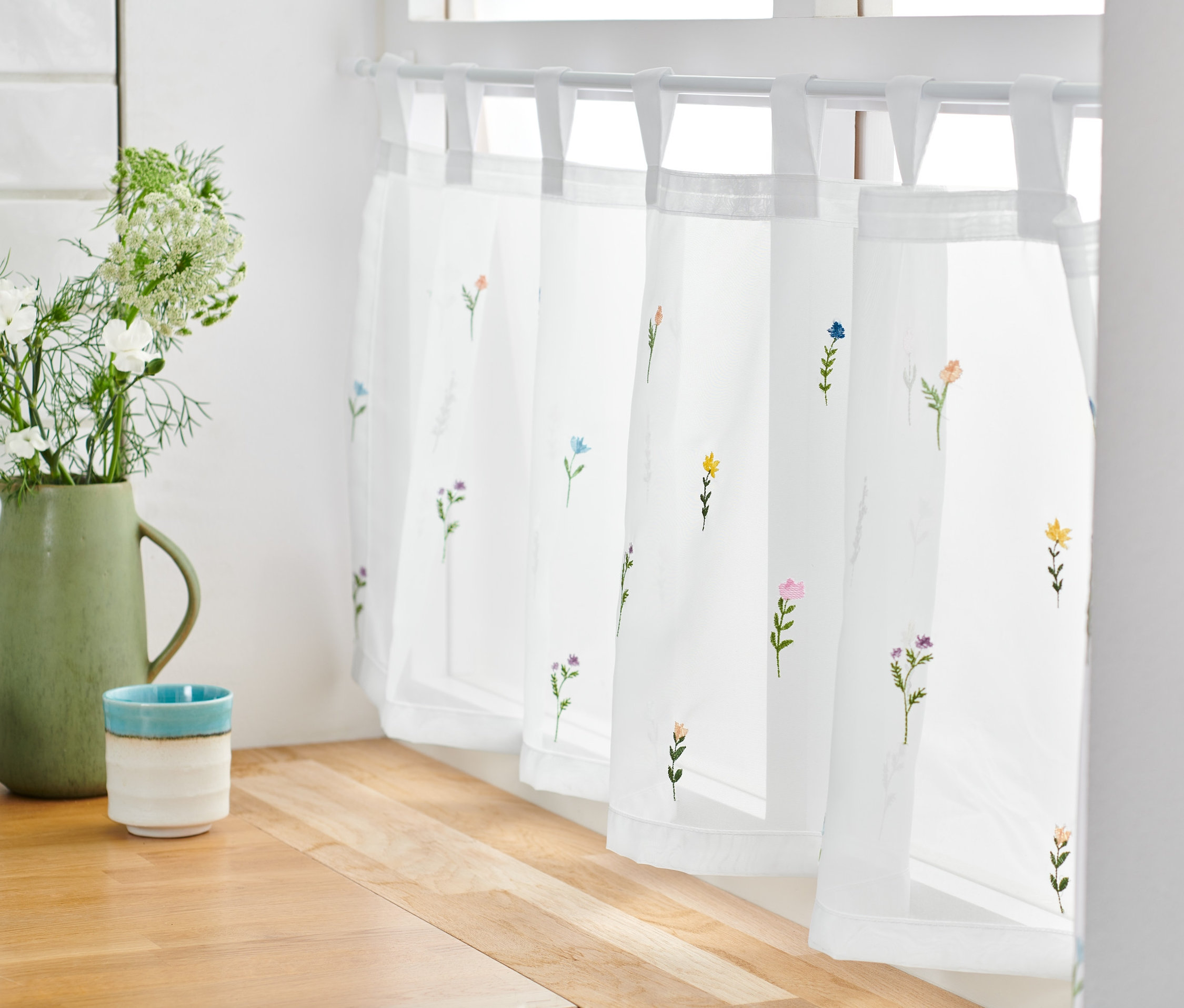 Bílá vyšívaná krátká záclona s květinovým motivem visí v okně.