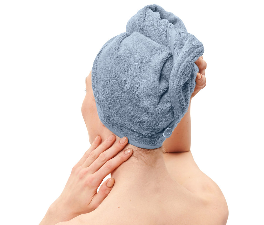 Žena s ručníkovým turbanem světle modré barvy na hlavě a ručníkem stejné barvy ovinutým kolem těla.