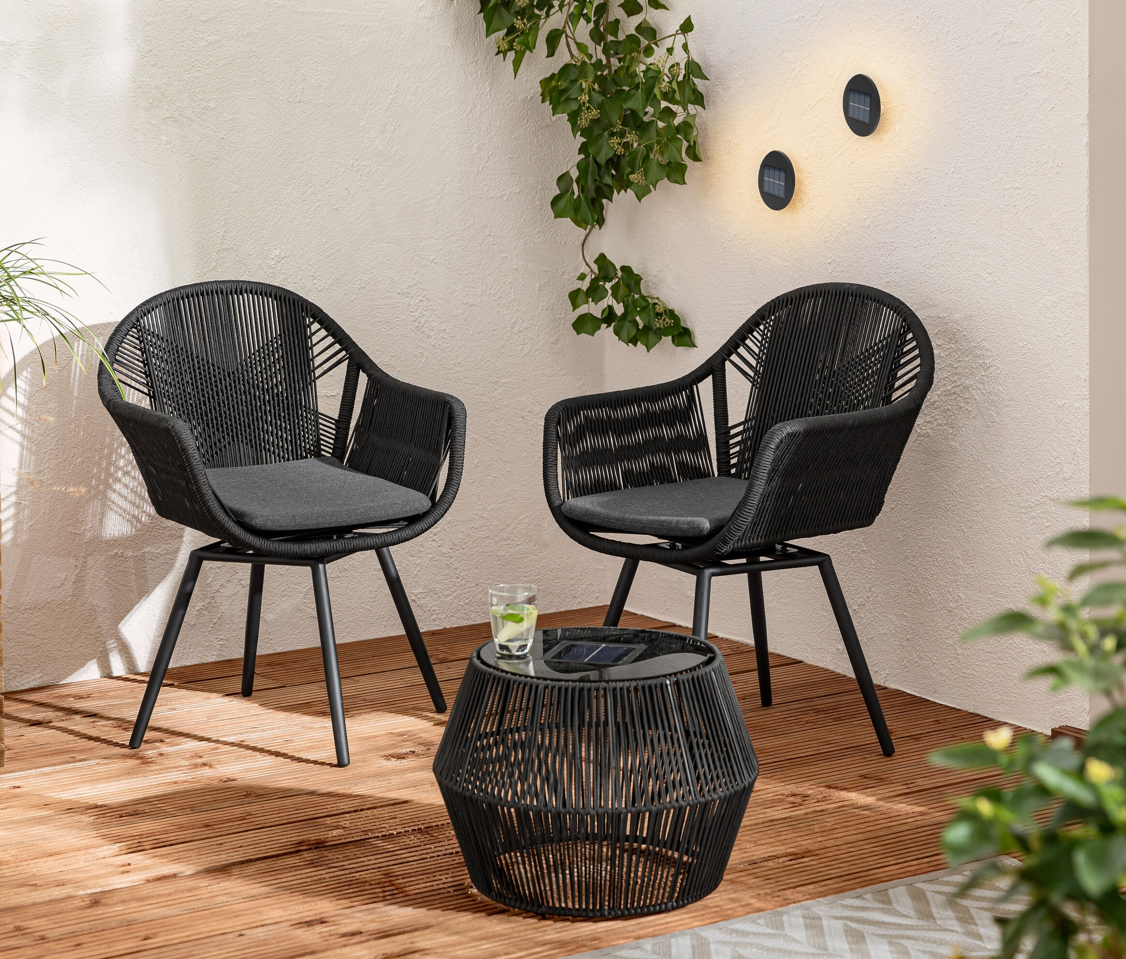 Dvě otočná jídelní křesla s textilním výpletem a solární odkládací stolek s textilním výpletem na dřevěné terase. Na zdi jsou 2 solární nástěnná a schodišťová svítidla.