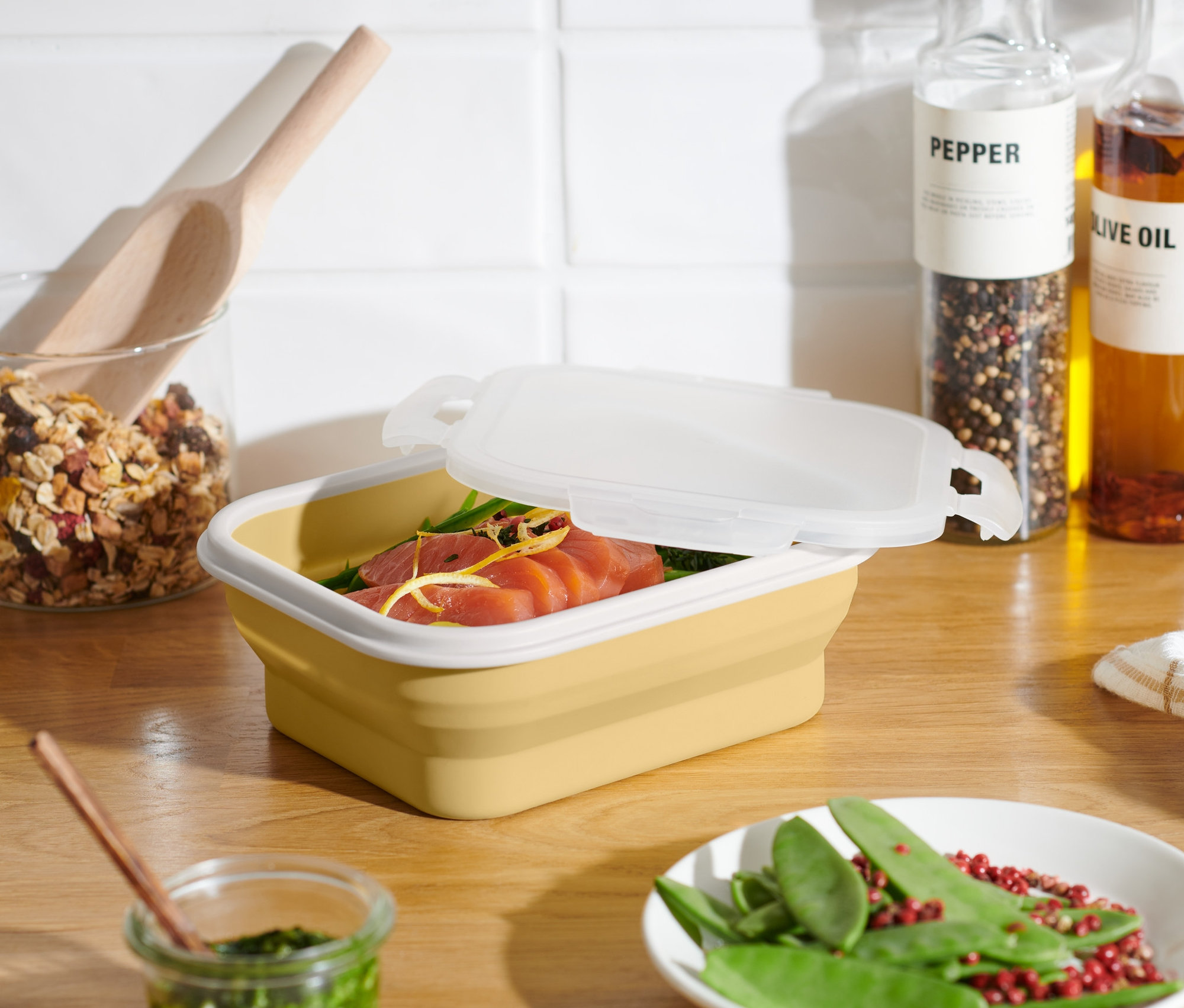 Žlutá skládací dóza na potraviny s pootevřeným víkem, uvnitř je losos. Na stole leží talíř se zeleným hráškem a červeným pepřem.