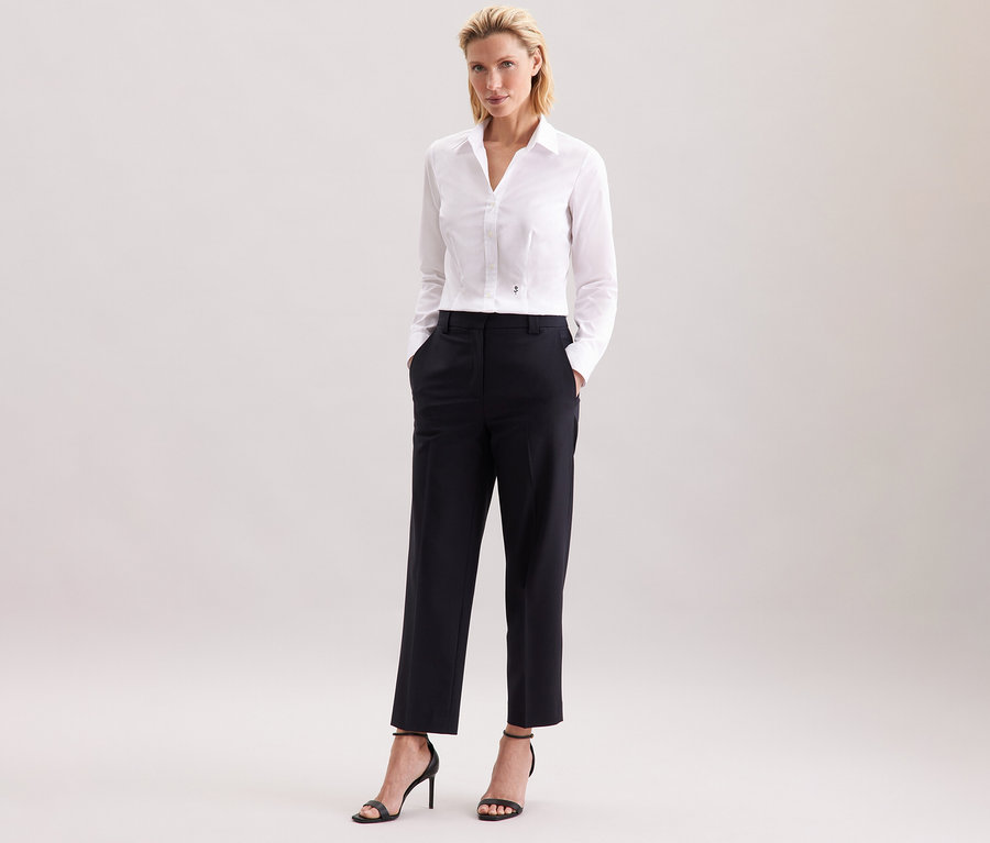 Žena stojí a má na sobě bílou halenku Seidensticker Slim Fit non iron, černé kalhoty a černé vysoké podpatky.