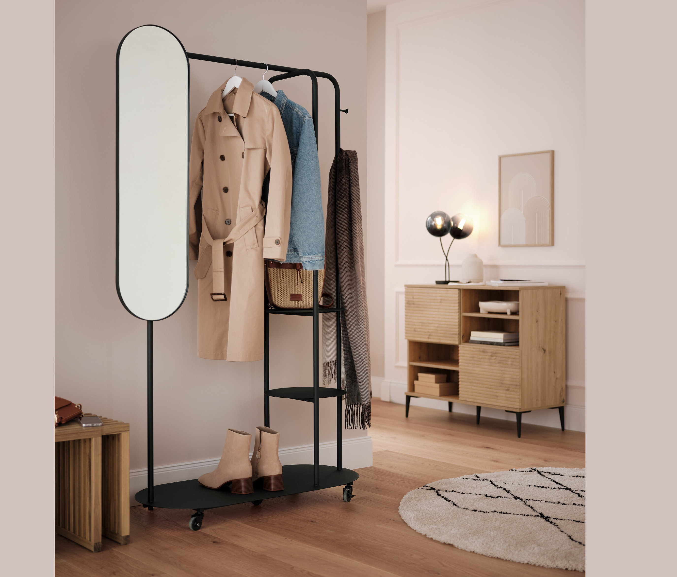 Černý věšák na oblečení s oválným zrcadlem a rozkládací stoličkou. Na věšáku visí béžový kabát, modrá bunda a šedá šála.