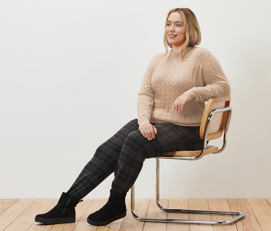Žena sedí na židli a má na sobě krémový svetr s copánkovým vzorem a kostkované kalhoty joggpant.