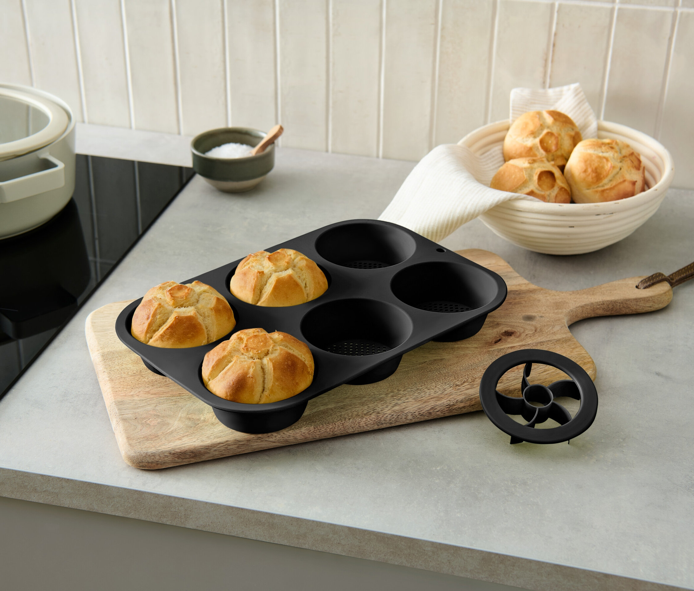 Tři upečené bulky v černé formě na pečení na dřevěném prkénku vedle mísy s bulkami.