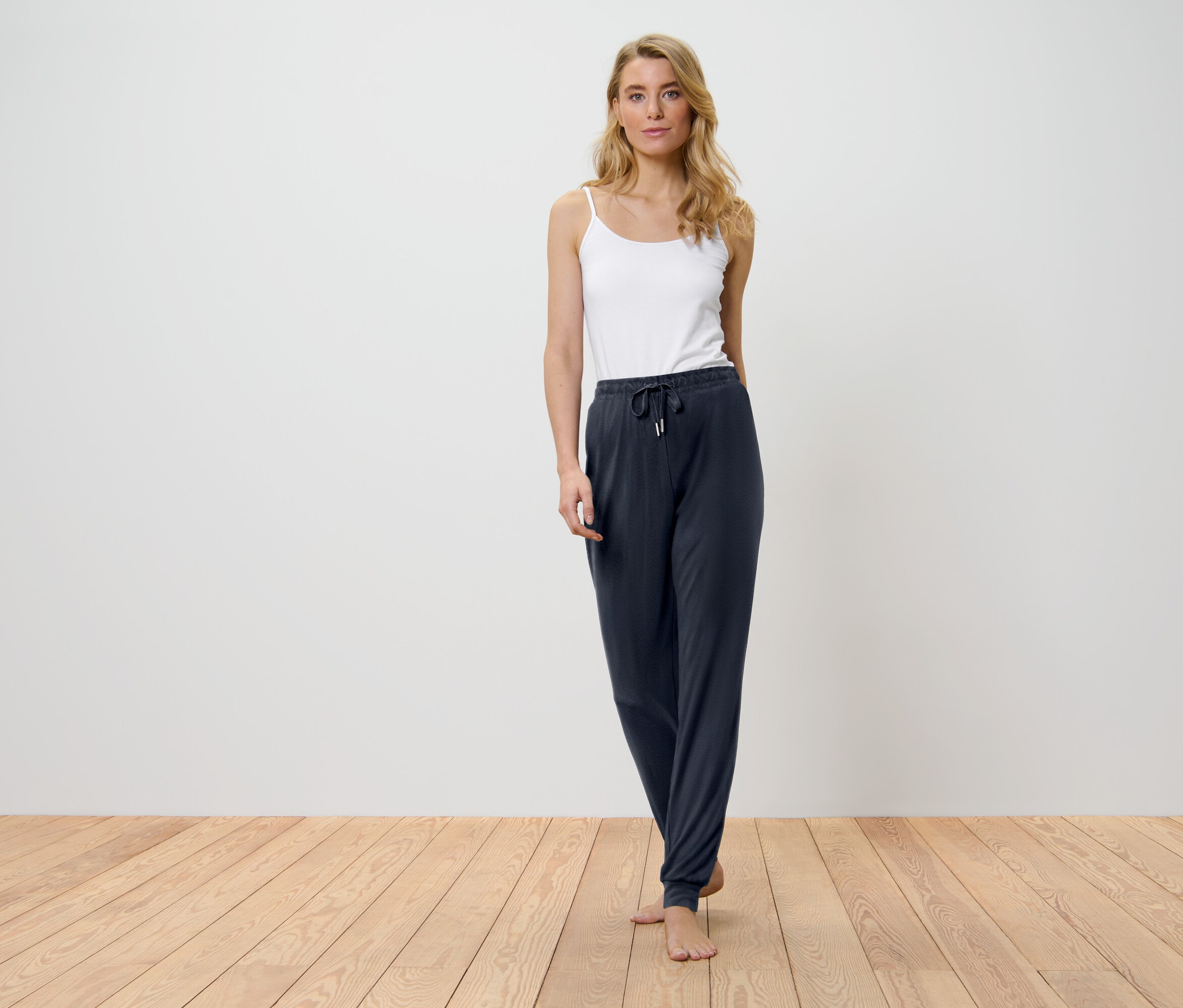 Blonďatá žena stojí na dřevěné podlaze a má na sobě bílý top a tmavě modré kalhoty.