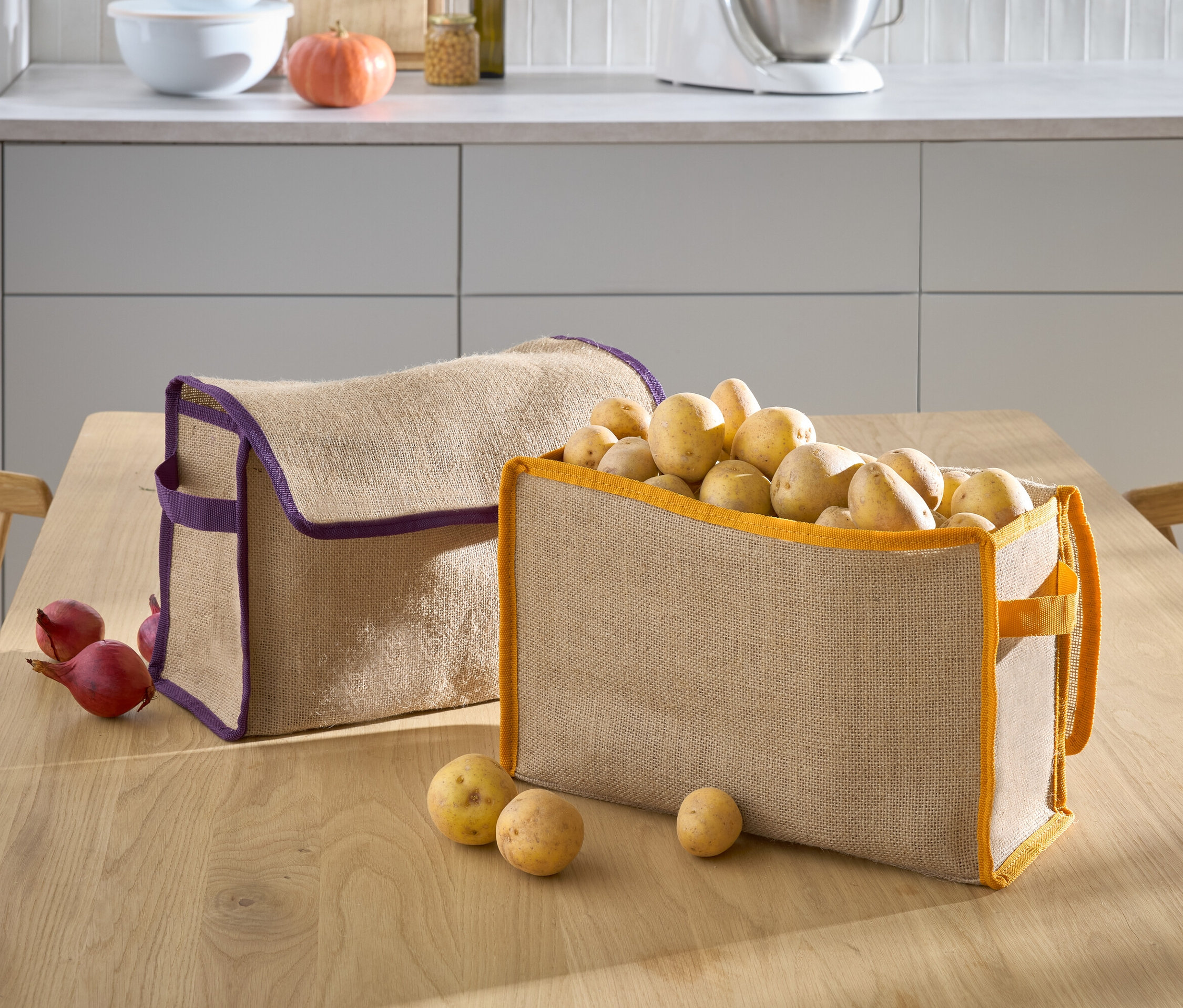 Dva béžové košíky leží na stole, jeden s fialovým lemem je prázdný a druhý s oranžovým lemem je plný brambor.