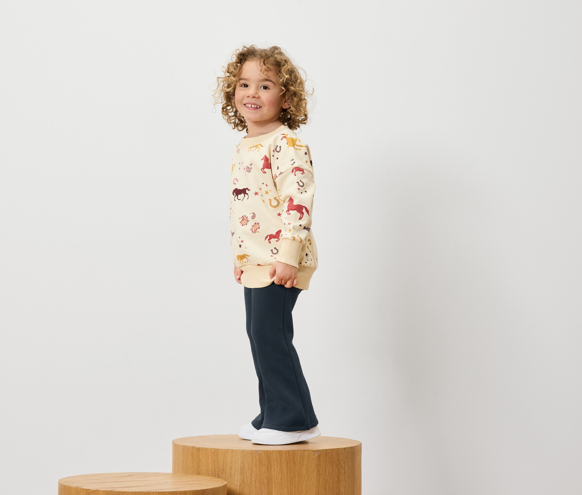 Chlapec s kudrnatými vlasy v mikině s potiskem koní a tmavě modrých kalhotách stojí na dřevěném podstavci.