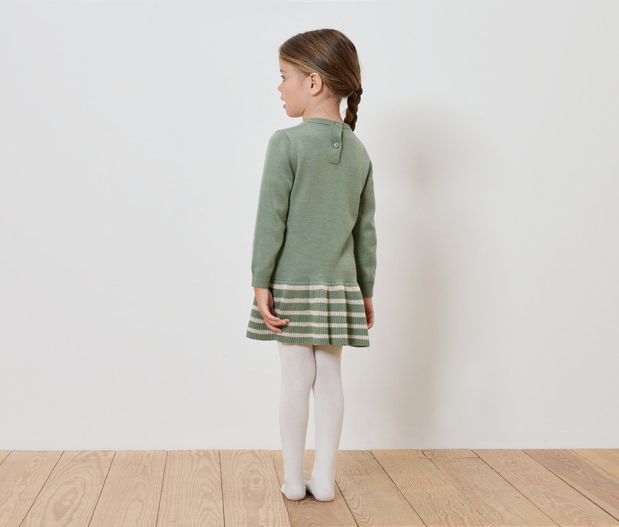 Dívka v zelených pletených šatech stojí zády ke kameře.