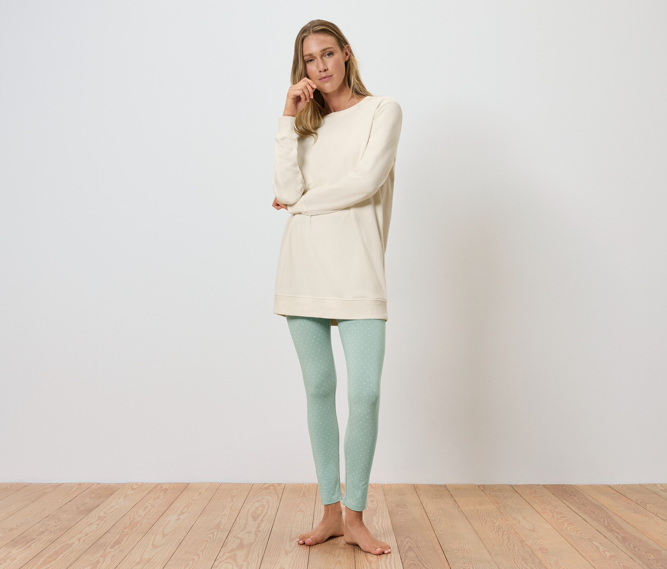Žena stojí a pózuje v krémové tunice a světle zelených legínách.