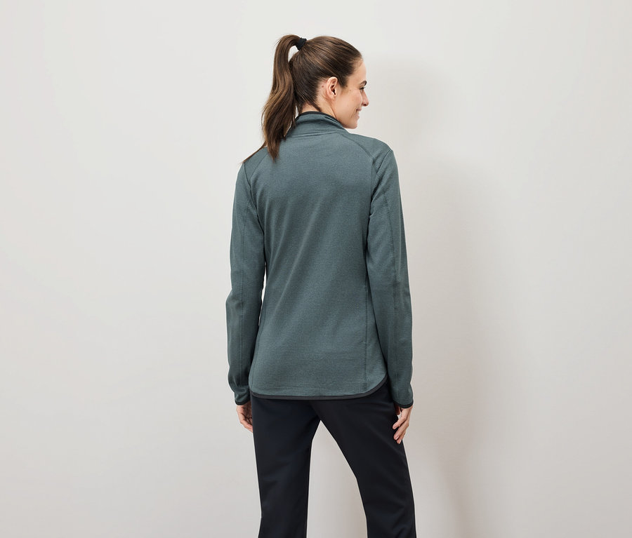 Zezadu je vidět žena s hnědými vlasy staženými do culíku, která má na sobě zelenou mikinu na zip a černé kalhoty.