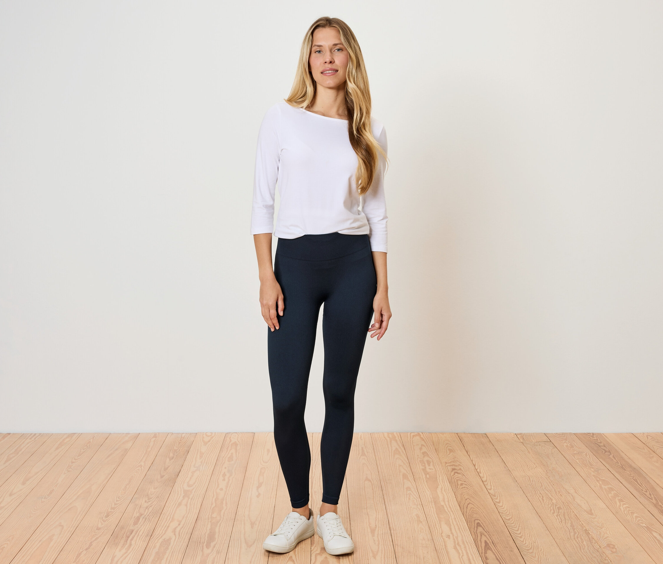 Žena stojí na dřevěné podlaze a má na sobě bílou košili s dlouhým rukávem a černé legíny.