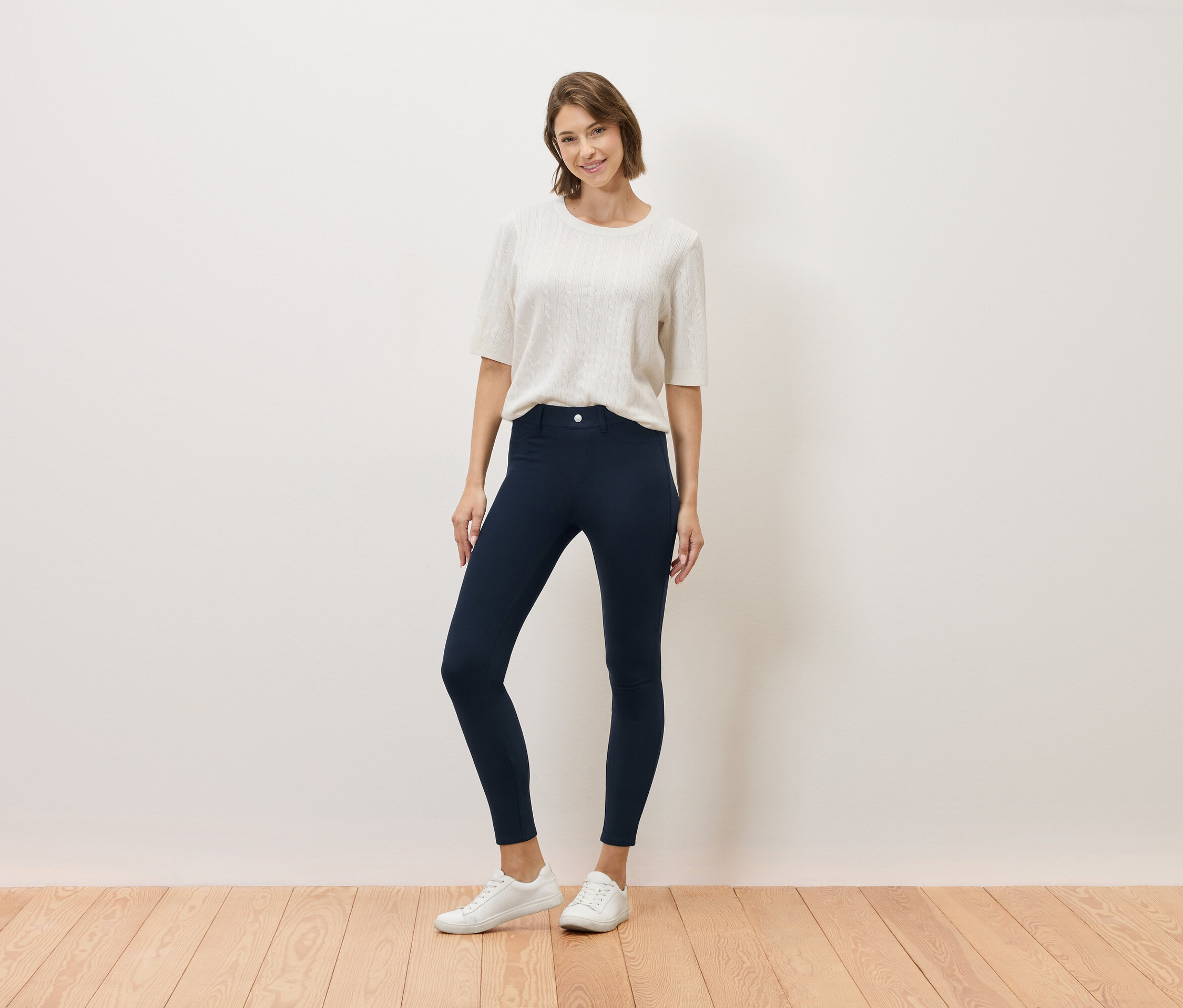 Žena stojí a pózuje v krémovém topu, tmavě modrých kalhotách a bílých teniskách.