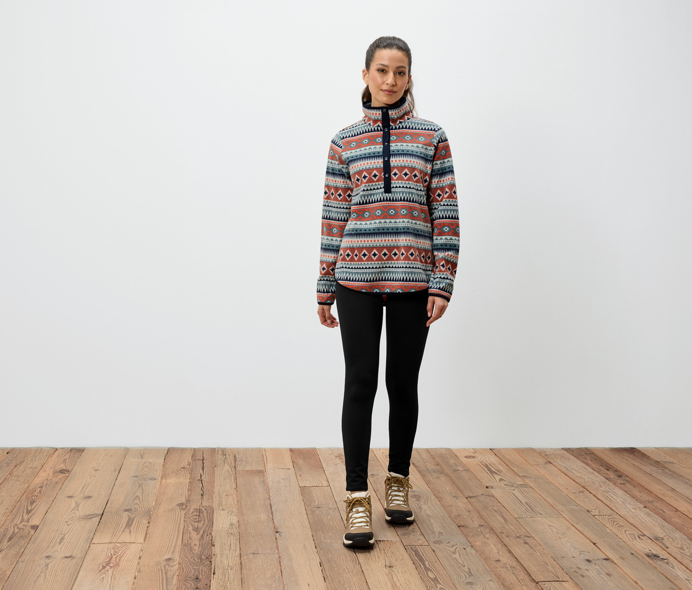 Žena stojí na dřevěné podlaze a má na sobě vzorovanou mikinu, černé legíny a hnědé boty.