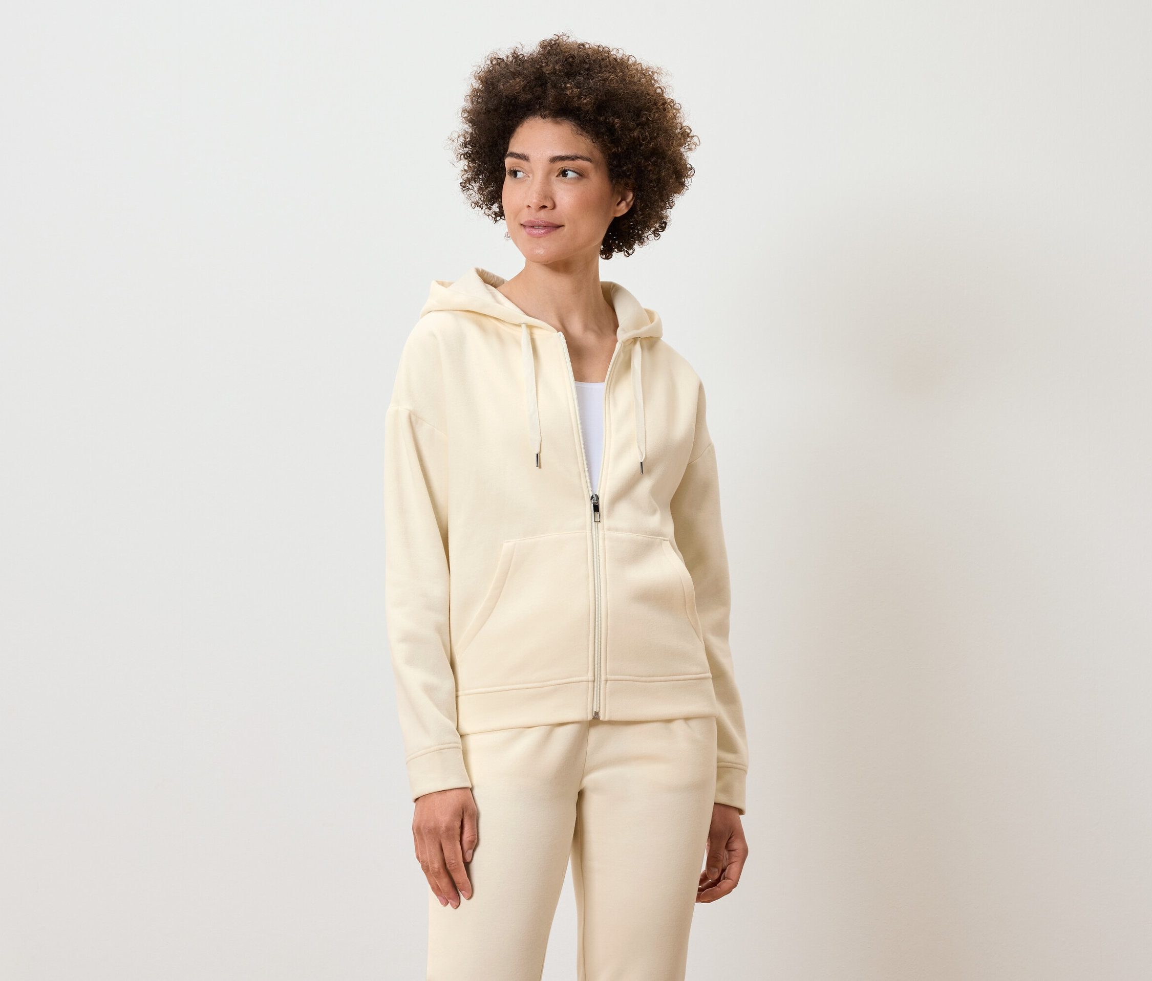 Žena s kudrnatými vlasy pózuje v krémově zbarvené mikině na zip s kapucí a teplákách.