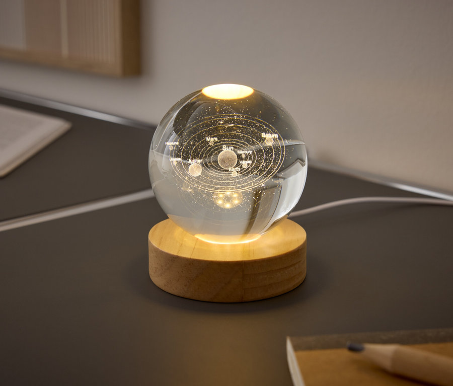 Skleněná koule s planetami sluneční soustavy na dřevěném podstavci svítí na stole.