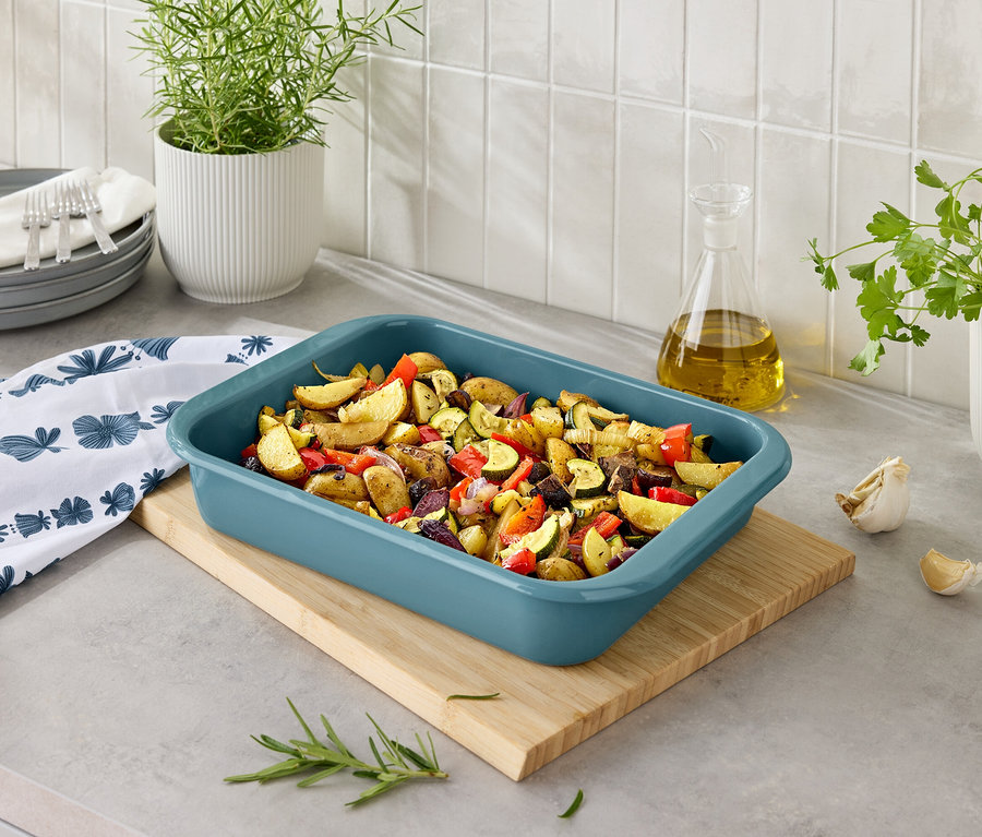 Pečené brambory se zeleninou v modré nádobě na pečení na dřevěném prkénku.