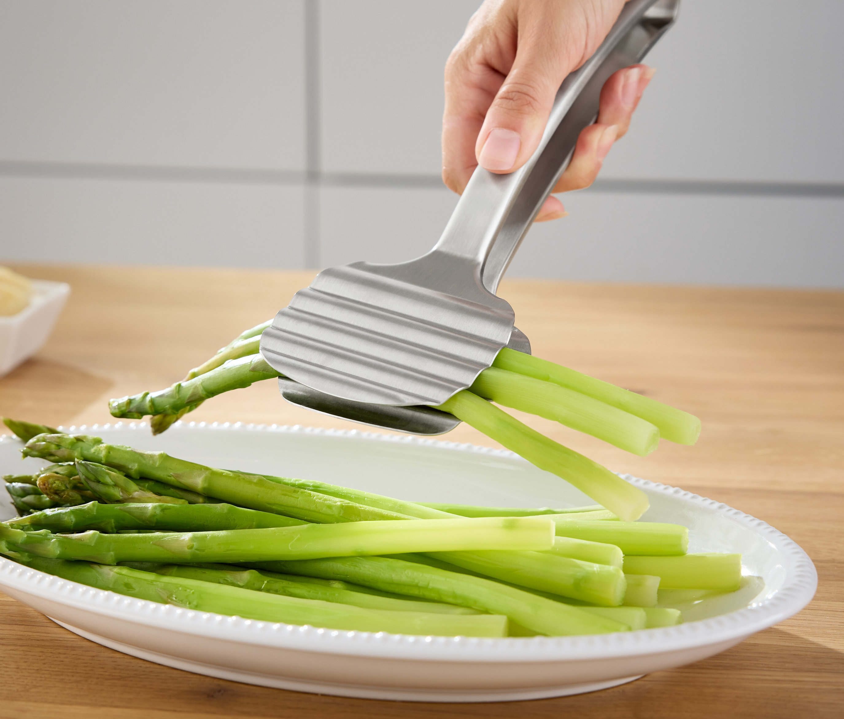 Ruka drží stříbrné kleště na chřest nad mísou se zeleným chřestem.