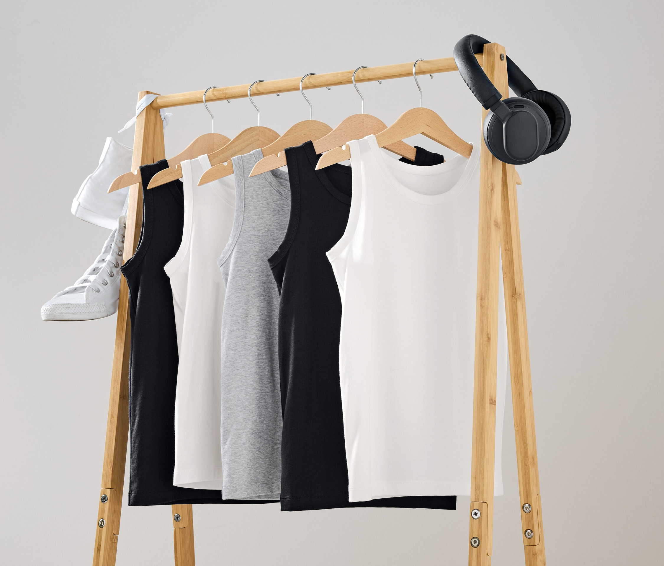 Na dřevěném věšáku visí tílka v černé, bílé a šedé barvě. Na věšáku je zavěšená bílá bota a černá sluchátka.