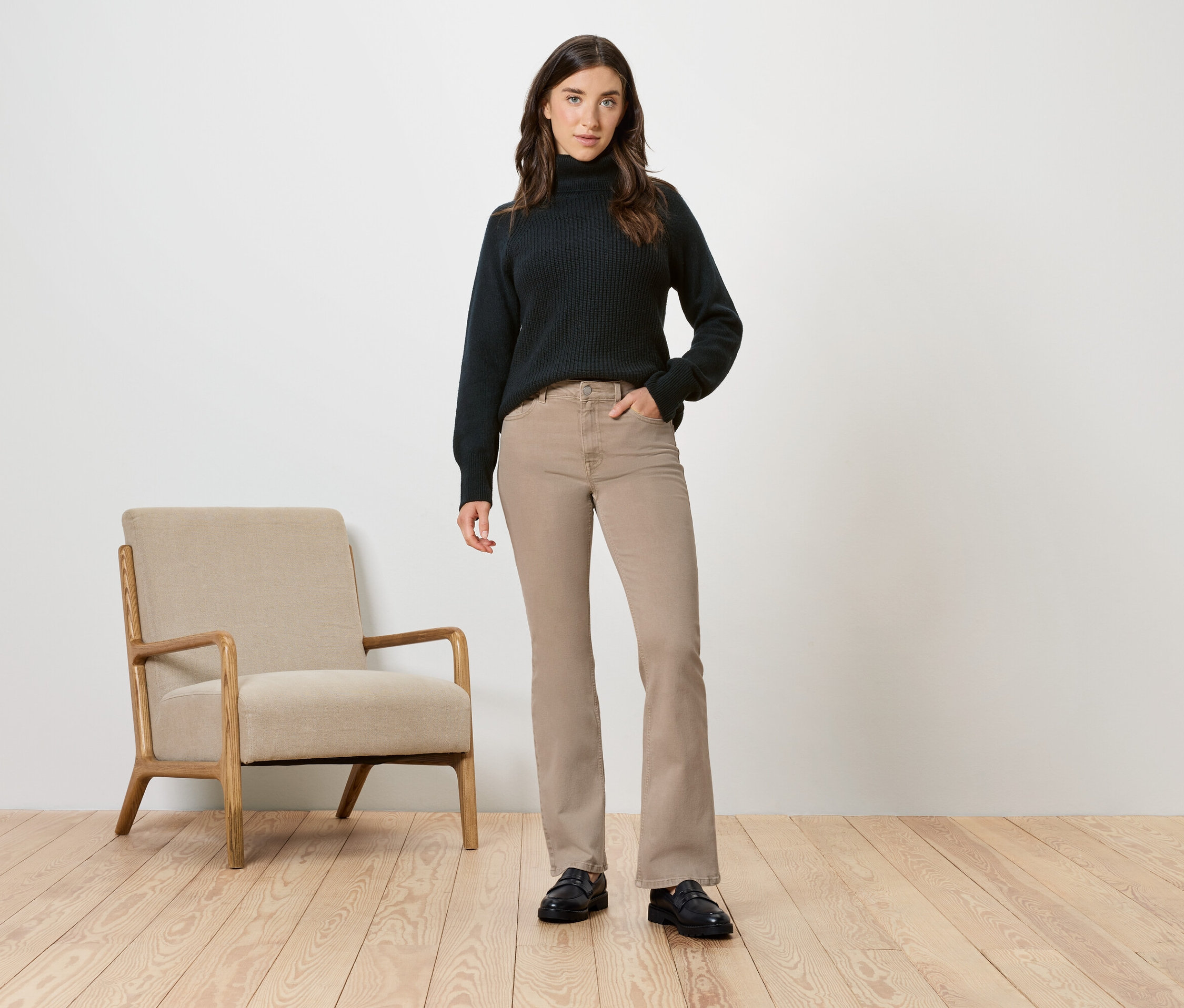 Žena v černém roláku a béžových kalhotách pózuje vedle béžového křesla.